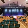 A school stage can be seen with Stars hanging from the curtains, the stage is lit with spotlights, and rows of empty chairs sit infront waiting for parents.