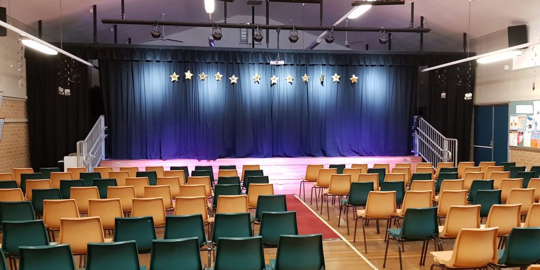 A school stage can be seen with Stars hanging from the curtains, the stage is lit with spotlights, and rows of empty chairs sit infront waiting for parents.