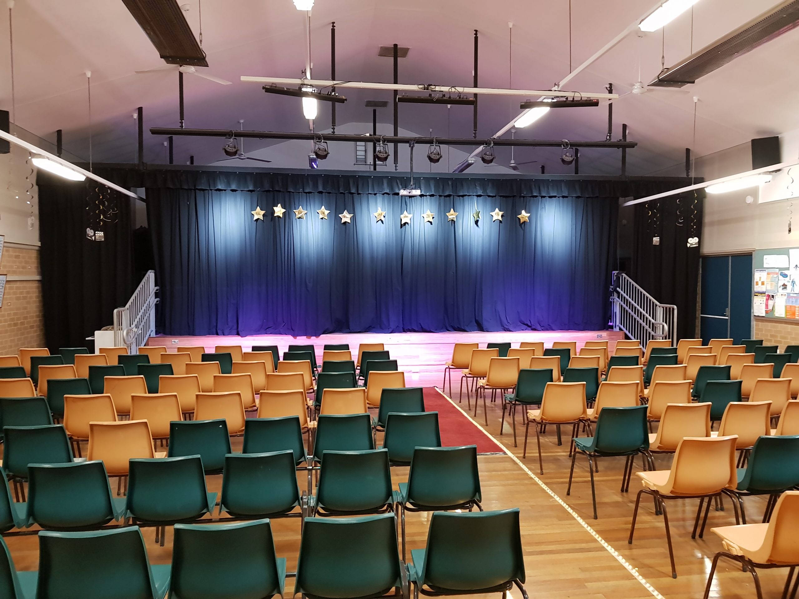 A school stage can be seen with Stars hanging from the curtains, the stage is lit with spotlights, and rows of empty chairs sit infront waiting for parents.