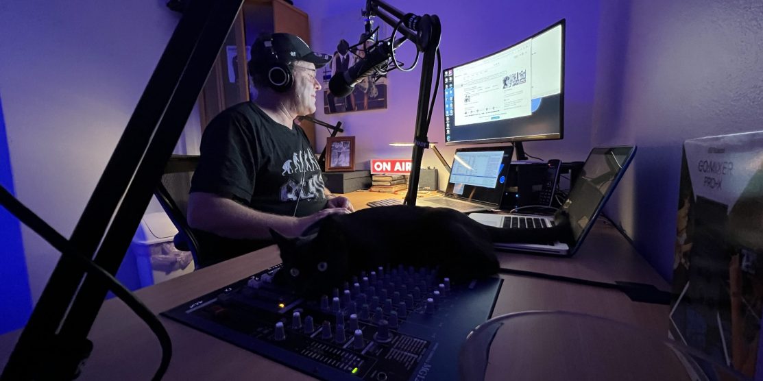 Paul Rothapfel sits at his desk in his studio. A cat is laying on the audio mixer controls.