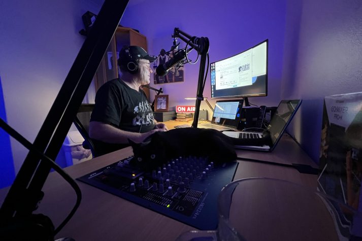 Paul Rothapfel sits at his desk in his studio. A cat is laying on the audio mixer controls.