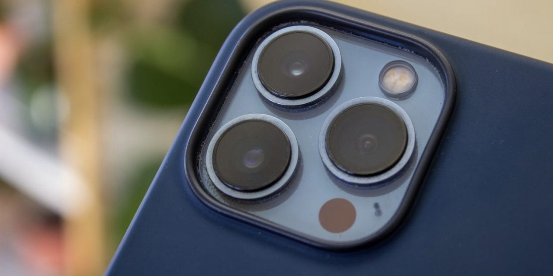 iPhone 13 Pro Max camera array with blue Magsafe case, blurred background with houseplants out of focus.