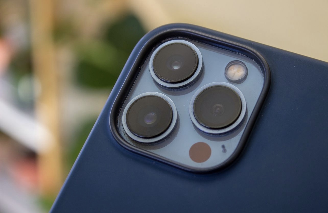 iPhone 13 Pro Max camera array with blue Magsafe case, blurred background with houseplants out of focus.