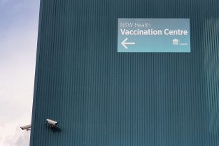 Green tin roof, and CCTV cameras site next to sign that reads 'NSW Health, Vaccination Centre.'