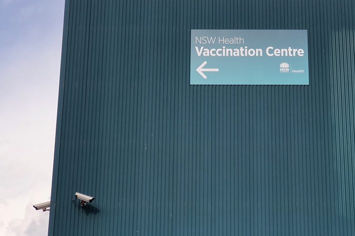 Green tin roof, and CCTV cameras site next to sign that reads 'NSW Health, Vaccination Centre.'