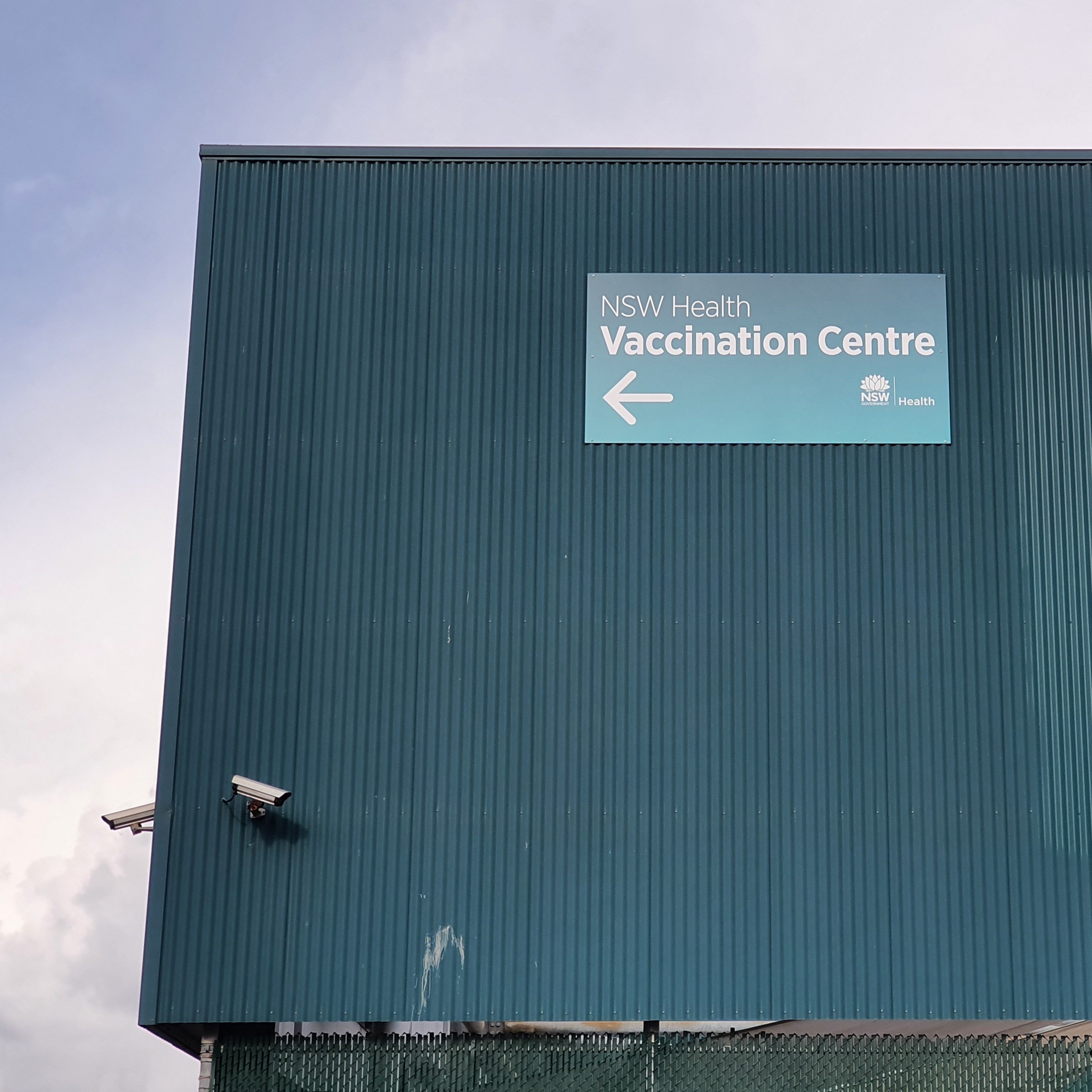 Green tin roof, and CCTV cameras site next to sign that reads 'NSW Health, Vaccination Centre.'